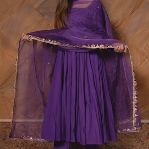 May include: A vibrant purple salwar kameez ensemble. The outfit includes a long, flowing tunic, matching pants, and a sheer dupatta with gold trim. The outfit is worn with gold-colored heels and large earrings. The background is a textured, brown wall.