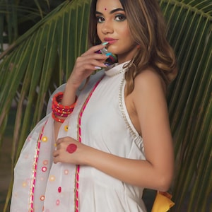 A white halter dress with gold trim and a matching sheer scarf. The scarf has a pink and gold border with colorful circular accents. The dress is paired with colorful bracelets and earrings.