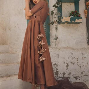 May include: A brown dress with short sleeves and a flowing skirt. The dress features decorative tassels and is paired with matching pants and gold heels. A brown scarf is draped on the ground.
