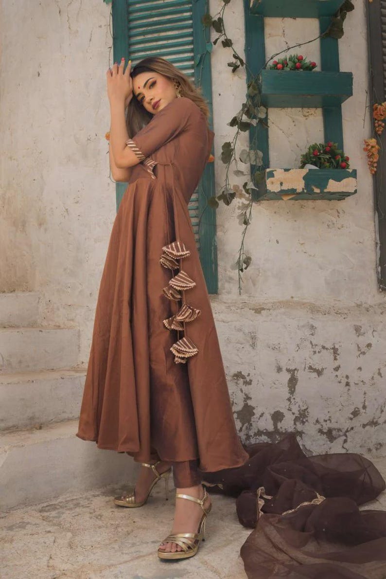 May include: A brown dress with short sleeves and a flowing skirt. The dress features decorative tassels and is paired with matching pants and gold heels. A brown scarf is draped on the ground.