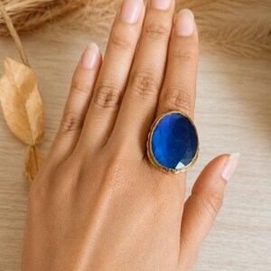 Könnte beinhalten: Ein Ring mit einem großen, ovalen, facettierten blauen Stein, der in eine strukturierte, goldfarbene Metallschiene gefasst ist. Der Stein zeigt eine tiefe, durchscheinende blaue Farbe mit sichtbaren Facetten, während die Fassung ein rustikales Finish aufweist.