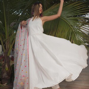 A flowing white dress with a halter neckline and a matching sheer scarf. The scarf is adorned with colorful floral embroidery and a pink border. The outfit is paired with gold heels.