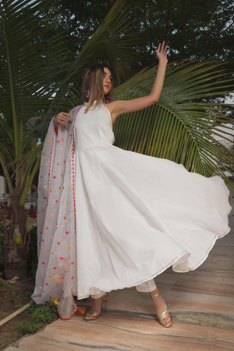 A flowing white dress with a halter neckline and a matching sheer scarf. The scarf is adorned with colorful floral embroidery and a pink border. The outfit is paired with gold heels.