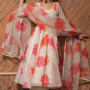 May include: A cream-colored Anarkali dress featuring a vibrant pink and orange floral print, paired with matching trousers and a sheer, lightweight dupatta with gold trim. The dress has long sleeves, a round neckline, and a flared silhouette.