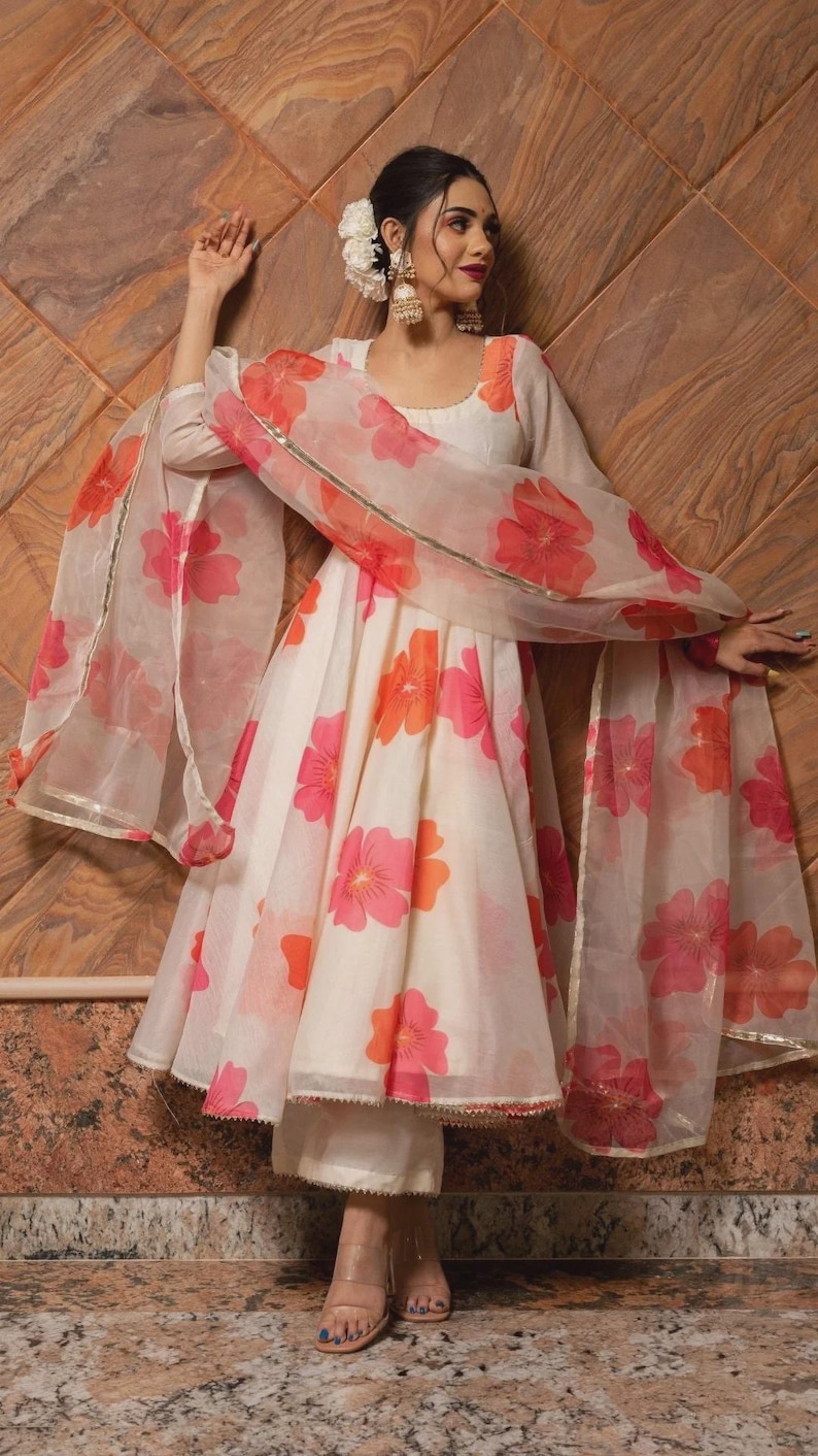 May include: A cream-colored Anarkali dress featuring a vibrant pink and orange floral print, paired with matching trousers and a sheer, lightweight dupatta with gold trim. The dress has long sleeves, a round neckline, and a flared silhouette.