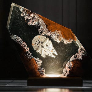 May include: A decorative lamp featuring a silver Millennium Falcon model encased in clear resin. The resin is shaped like a slanted rectangle, with brown and white rock-like formations on the sides. The lamp sits on a wooden base and emits a soft glow.
