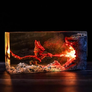 May include: A rectangular resin sculpture featuring a red and orange dragon breathing fire. The dragon is positioned in front of a fiery cave, with a dark background. The sculpture is illuminated from within, creating a dramatic effect.