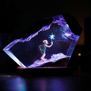 May include: A clear resin sculpture featuring a small, pale, large-eared creature wearing a green tunic, standing on a textured purple base. The creature holds a glowing blue star-shaped light, with wisps of smoke inside the resin enclosure.
