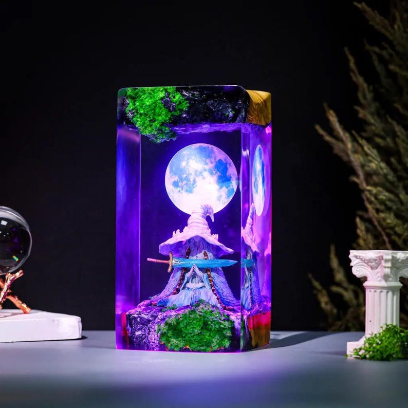 Pode incluir: Uma pe&ccedil;a de arte retangular em resina com uma cena de fantasia em miniatura. Apresenta uma lua cheia, um chap&eacute;u de bruxa, uma espada e uma montanha, iluminadas com luz roxa e azul. A parte superior tem detalhes verdes e pretos.