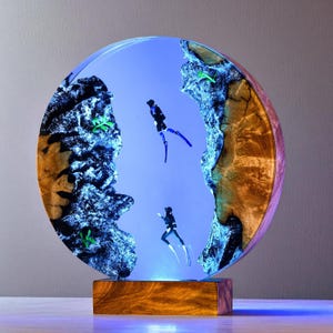 May include: A round resin sculpture featuring two scuba divers in a blue underwater scene. The sculpture has a wooden base and a blue light illuminating the scene. The edges of the sculpture are made of wood and resin.