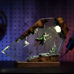 May include: A decorative resin lamp featuring a wizard on a broomstick, a castle, and an owl, set against a starry background. The lamp has a wooden base and a unique, geometric shape. The wizard is wearing a green cloak.