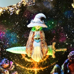 Pode incluir: Estatueta de uma bruxa com cabelo verde e chap&eacute;u branco, vestindo uma longa capa e segurando uma espada. A bruxa est&aacute; situada em um fundo de uma gal&aacute;xia colorida com estrelas.