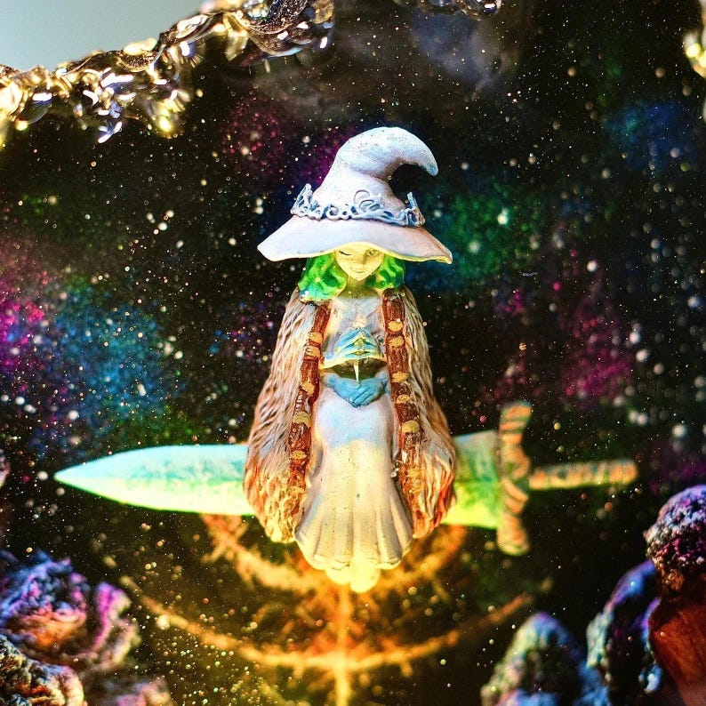 Pode incluir: Estatueta de uma bruxa com cabelo verde e chap&eacute;u branco, vestindo uma longa capa e segurando uma espada. A bruxa est&aacute; situada em um fundo de uma gal&aacute;xia colorida com estrelas.