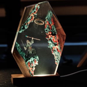 May include: A unique lamp featuring a fantasy scene encased in clear resin. The design includes a figure with a staff and sword, set against a backdrop of green and red rock formations. A gold ring floats above the figure. The lamp has a wooden base.