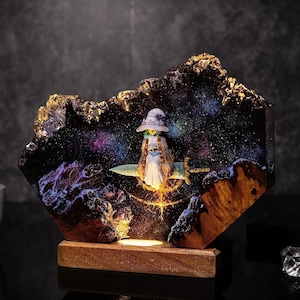 May include: A decorative resin lamp featuring a miniature figure of a witch riding a sword against a backdrop of a galaxy. The lamp has a wooden base and a dark, textured exterior with gold accents. The interior glows with colorful lights.