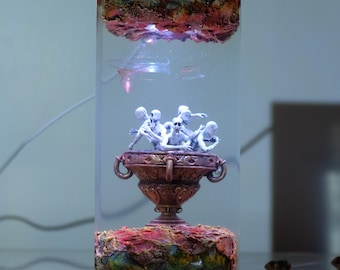May include: A rectangular resin sculpture featuring a decorative urn with several white skeleton figures. The sculpture has a layered design with red, brown, and green textured elements, and is illuminated with internal lighting.