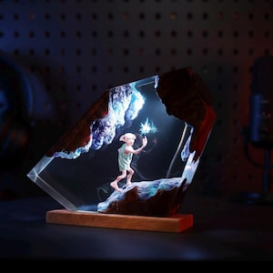 Puede incluir: Una escultura decorativa de resina que presenta un personaje con una túnica verde claro sosteniendo una estrella brillante. La escultura está encerrada en un bloque de resina transparente y angular con una base de madera y bordes marrones oscuros.