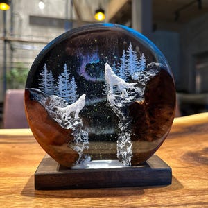 May include: A circular wooden sculpture depicting two white wolves howling under a night sky with an aurora borealis effect. The scene includes a forest, set within clear resin. The sculpture is mounted on a dark wooden base.