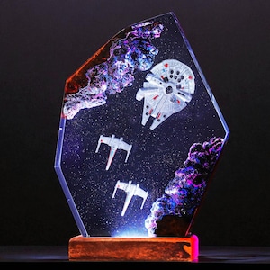 May include: A clear resin sculpture featuring a Millennium Falcon and two X-wing starfighters in a starry space scene. The artwork has purple and orange nebula-like accents and sits on a wooden base with a glowing pink light.
