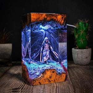 Puede incluir: Lámpara rectangular de resina con una figura encapuchada con dos sables de luz rojos. La lámpara tiene una base de madera y un interior azul y naranja brillante, creando un efecto de iluminación único. Un cactus decorativo está en el fondo.