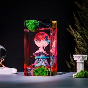 Pode incluir: Escultura retangular em resina com uma figura de mago em miniatura com chap&eacute;u rosa e espada azul, contra um fundo de lua vermelha e azul. A escultura tem detalhes em verde e preto, criando um tema de fantasia.