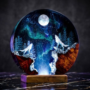 May include: A circular resin artwork showing a night scene with two white wolves howling at the moon. The piece includes a starry sky, aurora borealis, and a wooden base. The diameter is approximately 20 cm.