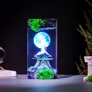 Pode incluir: Uma pe&ccedil;a de arte em resina retangular com uma cena de fantasia em miniatura. Inclui uma lua azul brilhante, uma pequena casa com um chap&eacute;u pontudo e folhagem verde. A obra de arte &eacute; iluminada com luz azul, criando um ambiente m&aacute;gico.