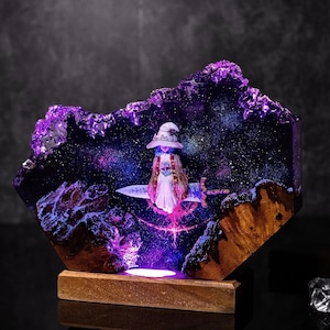 Pode incluir: Escultura em resina de uma bruxa montada numa espada atrav&eacute;s de uma gal&aacute;xia, com base em madeira e ilumina&ccedil;&atilde;o roxa e azul. A figura da bruxa est&aacute; vestida de branco. A escultura evoca um tema m&aacute;gico e de fantasia, com um fundo estrelado detalhado.