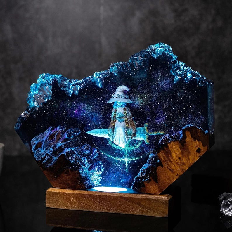 Pode incluir: Uma pe&ccedil;a de arte em resina &uacute;nica com uma cena celestial com uma figura em uma espada. A obra de arte tem uma base de madeira e um fundo azul com tema de gal&aacute;xia. A figura usa um chap&eacute;u e um vestido branco. A pe&ccedil;a &eacute; iluminada com luz azul.