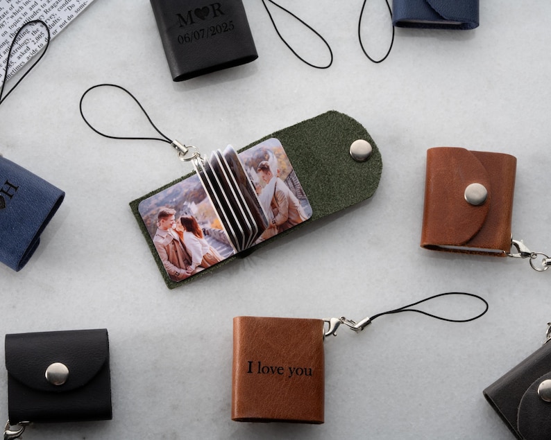 Op de afbeelding: Een verzameling kleine leren fotoalbums in verschillende kleuren, waaronder zwart, bruin en groen. Sommige albums zijn open en tonen afgedrukte foto's. E&eacute;n album is voorzien van de tekst "I love you".