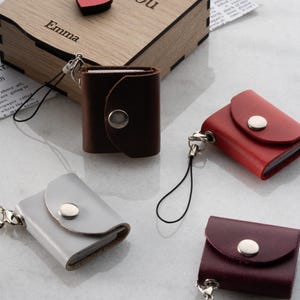 Op de afbeelding: Een verzameling kleine, leren notitieboekjes in verschillende kleuren: bruin, rood, grijs en bordeaux. Elk notitieboekje heeft een klepsluiting met een zilveren drukknop. Ze zijn bevestigd aan een zwart koord met een zilveren sluiting. Op de achtergrond staat een houten kist met de naam "Emma".