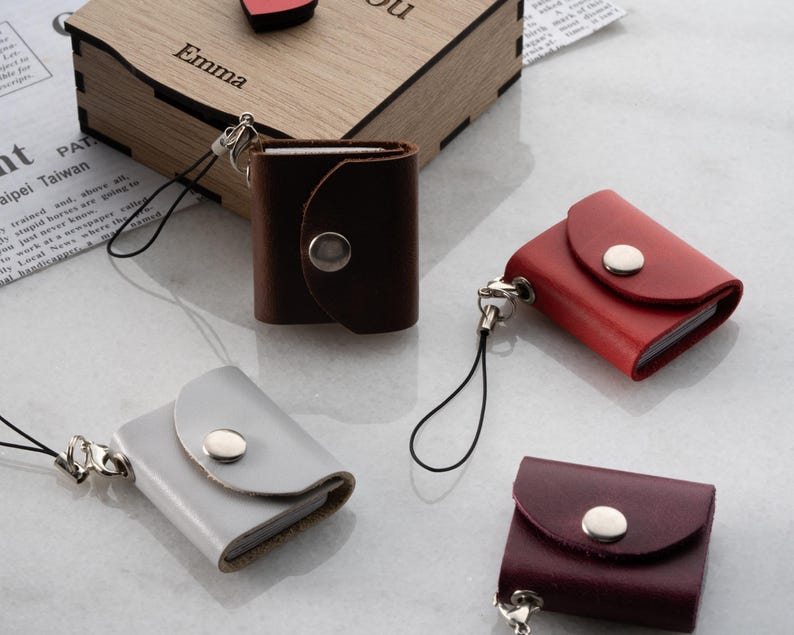 Op de afbeelding: Een verzameling kleine, leren notitieboekjes in verschillende kleuren: bruin, rood, grijs en bordeaux. Elk notitieboekje heeft een klepsluiting met een zilveren drukknop. Ze zijn bevestigd aan een zwart koord met een zilveren sluiting. Op de achtergrond staat een houten kist met de naam "Emma".