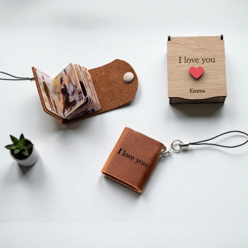 Op de afbeelding: Een verzameling gepersonaliseerde cadeaus, waaronder een klein leren fotoalbum, een houten doos met "I love you" en een rood hart, en een miniatuur leren boekje met "I love you" erop gedrukt. Er is ook een kleine potplant aanwezig.