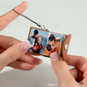 Op de afbeelding: Een kleine, bruine leren fotoalbum-bedel met een zilverkleurige sluiting en een zwart koord. Het album is open en toont kleurenfoto's. Het album wordt tussen twee handen gehouden.