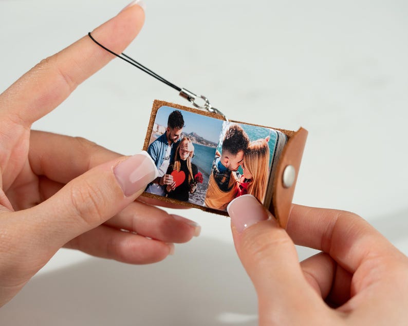 Op de afbeelding: Een kleine, bruine leren fotoalbum-bedel met een zilverkleurige sluiting en een zwart koord. Het album is open en toont kleurenfoto's. Het album wordt tussen twee handen gehouden.
