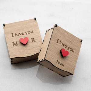 Op de afbeelding: Twee houten dozen met scharnierende deksels. Op elke doos staat de tekst "I love you" gegraveerd op het deksel, samen met een rood hart en initialen of een naam. De dozen zijn gemaakt van lichtgekleurd hout.