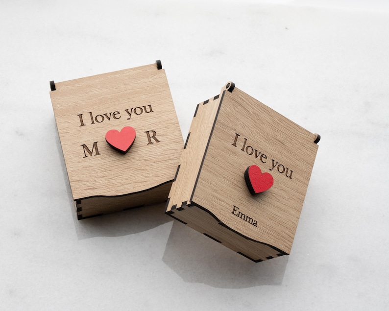 Op de afbeelding: Twee houten dozen met scharnierende deksels. Op elke doos staat de tekst "I love you" gegraveerd op het deksel, samen met een rood hart en initialen of een naam. De dozen zijn gemaakt van lichtgekleurd hout.