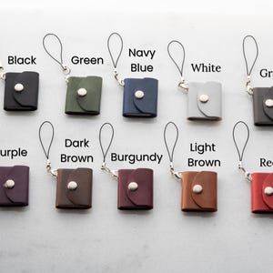 Op de afbeelding: Een verzameling kleine, vierkante leren etuis in verschillende kleuren, waaronder zwart, groen, marineblauw, wit, grijs, paars, donkerbruin, bordeauxrood, lichtbruin en rood. Elk etui heeft een zilveren knoopsluiting en een polsbandje.