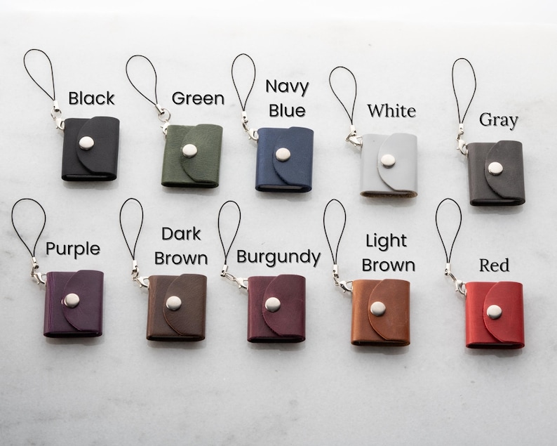 Op de afbeelding: Een verzameling kleine, vierkante leren etuis in verschillende kleuren, waaronder zwart, groen, marineblauw, wit, grijs, paars, donkerbruin, bordeauxrood, lichtbruin en rood. Elk etui heeft een zilveren knoopsluiting en een polsbandje.