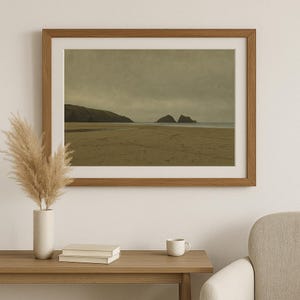 May include: A framed landscape print depicts a beach scene under a cloudy sky. The artwork features a sandy beach, rolling hills, and two rock formations in the distance. The frame is made of wood, and the overall color palette is muted with earthy tones.