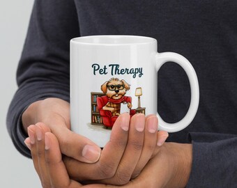 Pode incluir: Caneca de cerâmica branca com o texto "Pet Therapy". A caneca apresenta uma ilustração de desenho animado de um cão usando óculos e um cachecol, sentado em uma poltrona vermelha. Uma pequena lâmpada e uma estante também são retratadas.