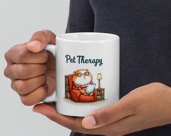 Pode incluir: Caneca de cerâmica branca com a frase "Pet Therapy". A caneca apresenta uma ilustração de um hamster com óculos e cachecol, lendo em uma poltrona vermelha. Uma pequena lâmpada e uma estante também são retratadas.