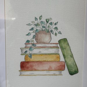 Op de afbeelding: Aquarel schilderij van een stapel boeken met een potplant erop. De boeken zijn in tinten rood, geel en groen. Een enkel groen boek leunt tegen de stapel. De plant heeft groene bladeren en een bruine pot. Het kunstwerk is wit omlijst.