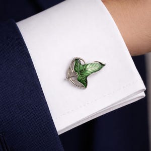 Op de afbeelding: Zilveren manchetknop met een groen bladerontwerp. Het blad heeft gedetailleerde textuur en verschillende groentinten. De manchetknop is geplaatst op een wit overhemd en een marineblauw pak.