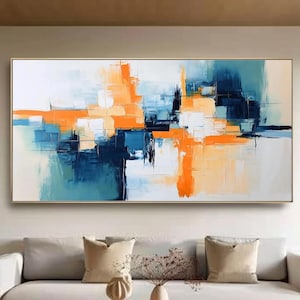 May include: Abstract art painting with a gold frame. The artwork features a vibrant composition of orange, blue, and white brushstrokes. The painting hangs above a white sofa in a living room setting, creating a modern and artistic ambiance.