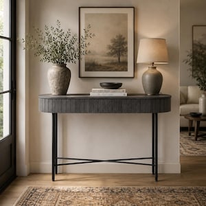 Op de afbeelding: Een donkergrijze ovale consoletafel met verticale latten en dunne zwarte metalen poten. Bovenop staat een gestructureerde stenen vaas met groene takken, een zwarte kom, twee gestapelde boeken met de titels Live Beautifully en Still Fashion, en een beige tafellamp.