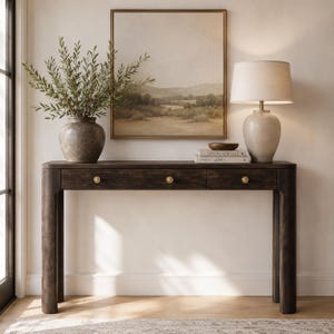 Peut inclure: Une console en bois brun foncé dotée de trois tiroirs, chacun muni d'un petit bouton rond en laiton. La table présente des bords arrondis et quatre pieds robustes. Un vase gris texturé avec de la verdure, une lampe et deux livres sont posés dessus.
