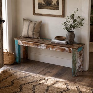 Op de afbeelding: Een rechthoekige houten bank met een verweerde afwerking in bruin, wit en turquoise. De bank heeft vier stevige poten en een vlakke zitting. Aan de linkerkant van de bank ligt een beige kussen met dunne, zwarte verticale strepen.