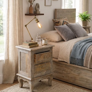 May include: A bedroom scene featuring a rustic wooden nightstand with two drawers, topped with books, a lamp, and a mug. A bed with pillows and a blue blanket is in the background. The room has a neutral color palette with natural light.