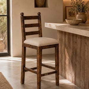 Puede incluir: Un taburete de bar alto hecho de madera oscura desgastada con un diseño de respaldo de escalera y dos remates redondeados en la parte superior. El asiento tiene un cojín de tela beige claro y la estructura incluye cuatro patas rectas.
